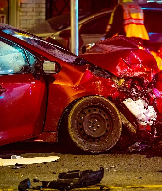 Red car with severe front-end damage at night, debris scattered on road, and emergency responder partially visible in background