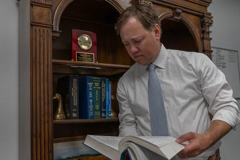 James E. Brogdon, III in white shirt and tie reading a large book in front of a wooden bookshelf with books and a clock trophy