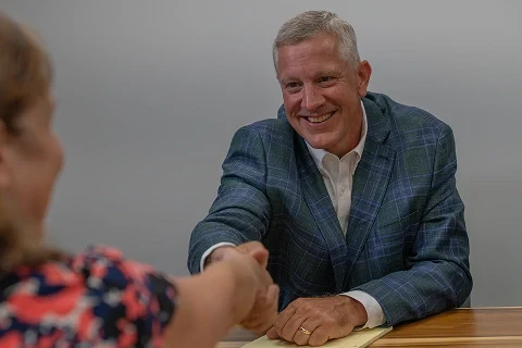 John Eric Fulda in a blue plaid blazer shakes hands with a woman across a wooden table in a professional setting