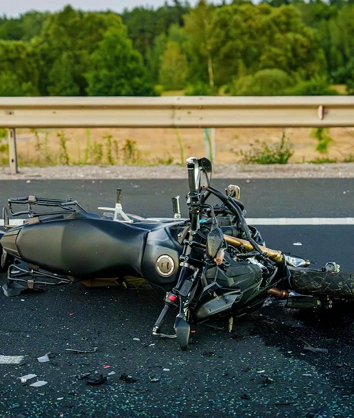 Black motorcycle lying on its side on a road, showing damage and debris, with a guardrail and trees in the background