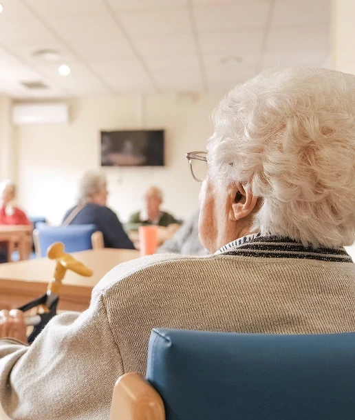 Elderly person with white hair sitting in a chair, holding a cane, in a room with other seniors in the background