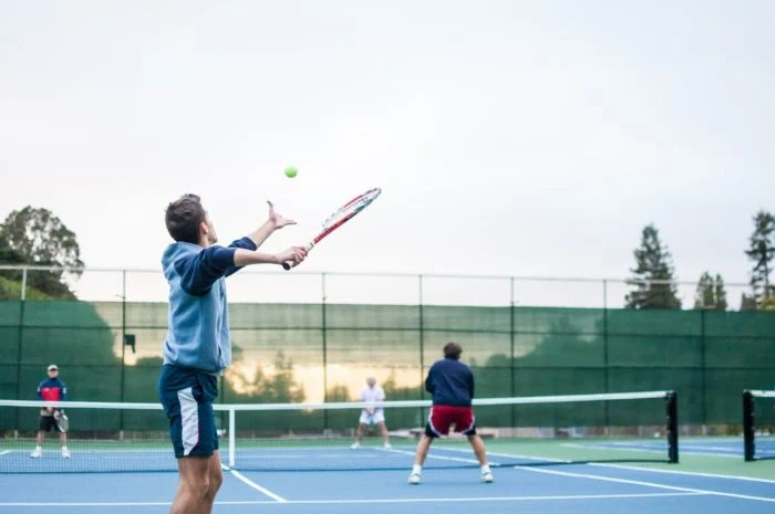 A man in a blue jacket serves a tennis ball on a court while three other players stand ready across the net
