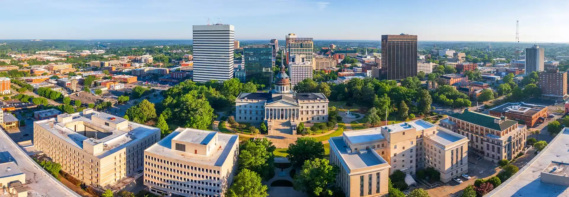 Aerial view of a city with a central historic capitol building surrounded by greenery and modern high-rise buildings on a clear day