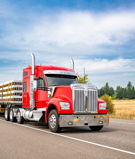 Red semi-truck hauling a flatbed trailer loaded with lumber, driving on a highway beside a field under a partly cloudy sky