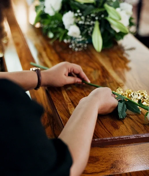 Person placing a single white rose on a polished wooden casket during a funeral service, with floral arrangements visible in the background